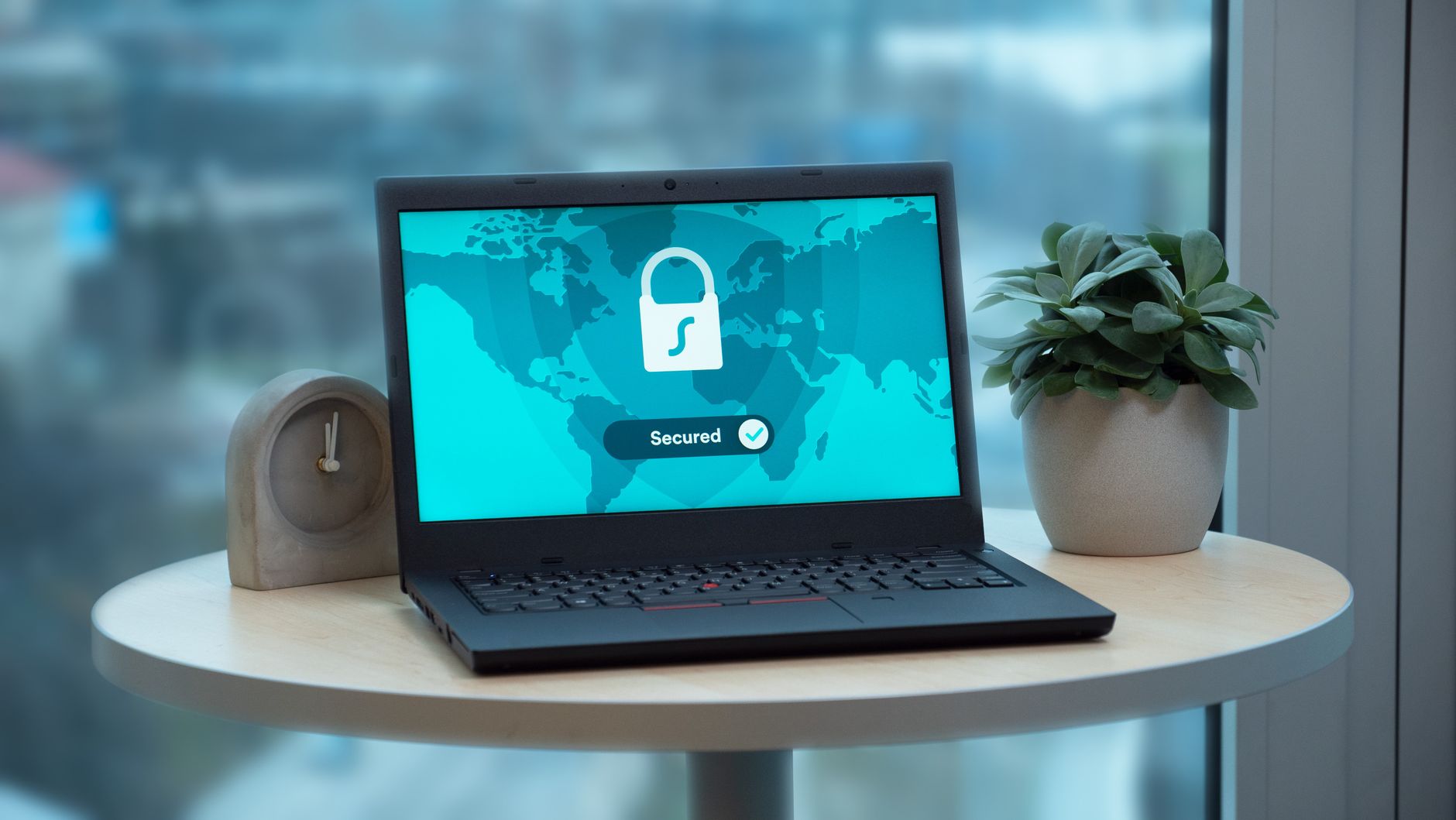 Laptop z ikoną kłódki na biurku obok doniczki i zegara